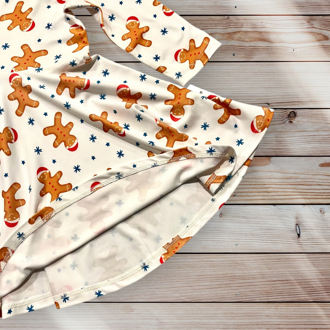 A long-sleeved, cream-colored dress for little girls age 12 months to 10 years old featuring a repeating pattern of gingerbread men wearing red hats and blue stars. The dress has a flared skirt and is designed for children. Festive and holiday-themed. Christmas dress