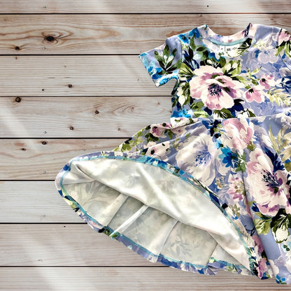 A light blue short-sleeved dress for little girls with a floral print featuring pink, white, blue, and green flowers. The dress has a full twirling skirt and a round neckline. size for ages 12 months to 12 years old. 