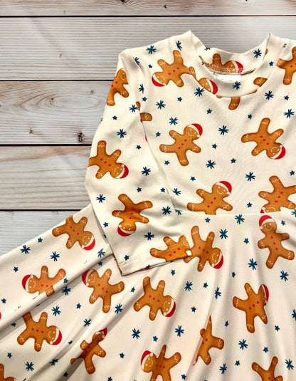 A long-sleeved, cream-colored dress for little girls age 12 months to 10 years old featuring a repeating pattern of gingerbread men wearing red hats and blue stars. The dress has a flared skirt and is designed for children. Festive and holiday-themed. Christmas dress