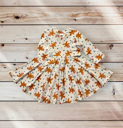 A long-sleeved, cream-colored dress for little girls age 12 months to 10 years old featuring a repeating pattern of gingerbread men wearing red hats and blue stars. The dress has a flared skirt and is designed for children. Festive and holiday-themed. Christmas dress